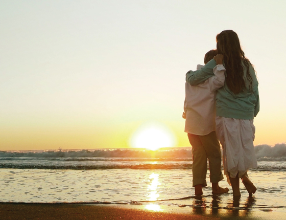 A young woman and a young boy stand on the beach looking at the setting sun with their backs to the camera